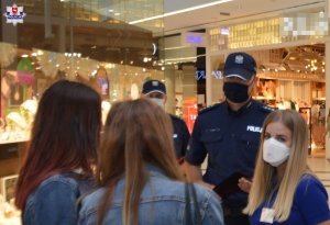 Umundurowany policjant w maseczce ochronnej stoi w grupie osób.