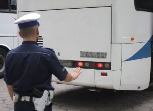 Umundurowany policjant w białej czapce stoi tyłem. Z przodu widoczny jest autobus.