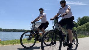 Umundurowani dwaj policjanci jadący na rowerach, patrolują bezpieczeństwo osób wypoczywających nad wodą