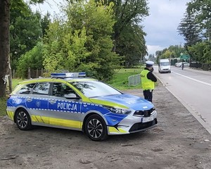 Zdjęcie przedstawia umundurowanego policjanta drogówki, który stając przy radiowozie oznakowanym kontroluje pomiar prędkości jadących pojazdów.