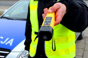 urządzenie do badania stanu trzeźwości trzymane w dłoni, w tle stoi radiowóz policyjny.