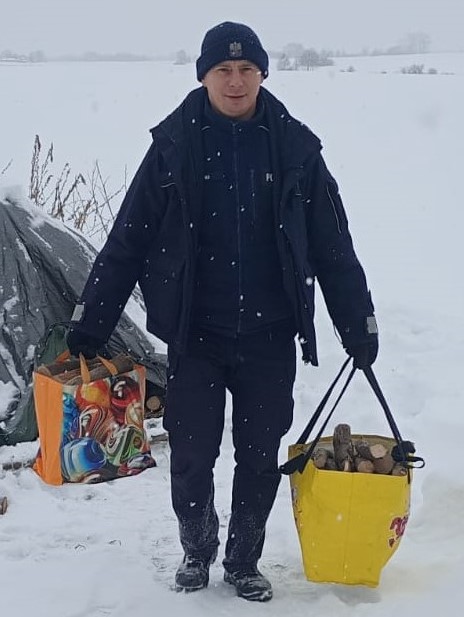 umundurowany policjant idzie przez zaśnieżoną drogę. W rękach trzyma torby z drewnem na opał
