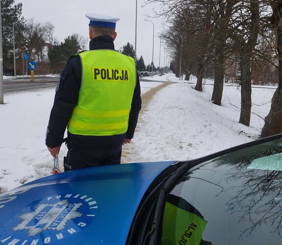 Na zdjęciu uwidoczniono umundurowanego policjanta drogówki, który stoi przy oznakowanym radiowozie